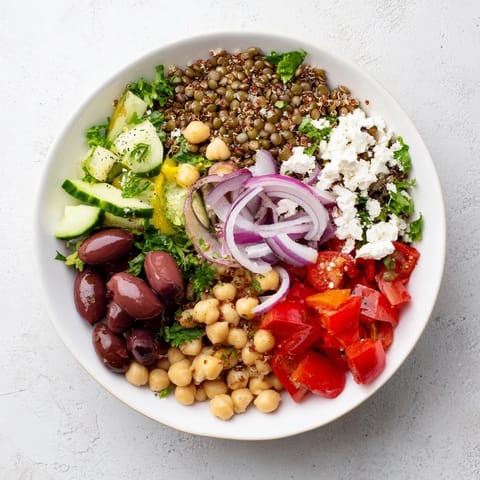 A vibrant Greek Power Salad in a white bowl, featuring quinoa, lentils, chickpeas, and fresh vegetables drizzled with zesty dressing.