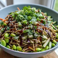 A close-up of a vibrant Soba Noodle Bowl topped with edamame, julienned carrots, cucumber, and scallions, drizzled with creamy sesame dressing.