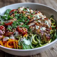 A vibrant Spiralized Vegetable Bowl with zucchini noodles, roasted sweet potato, cherry tomatoes, and grilled chicken drizzled with creamy tahini sauce.