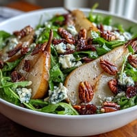 Fresh peppery arugula topped with sliced ripe pears, crumbled blue cheese, and toasted walnuts, drizzled with balsamic dressing in a white bowl.
